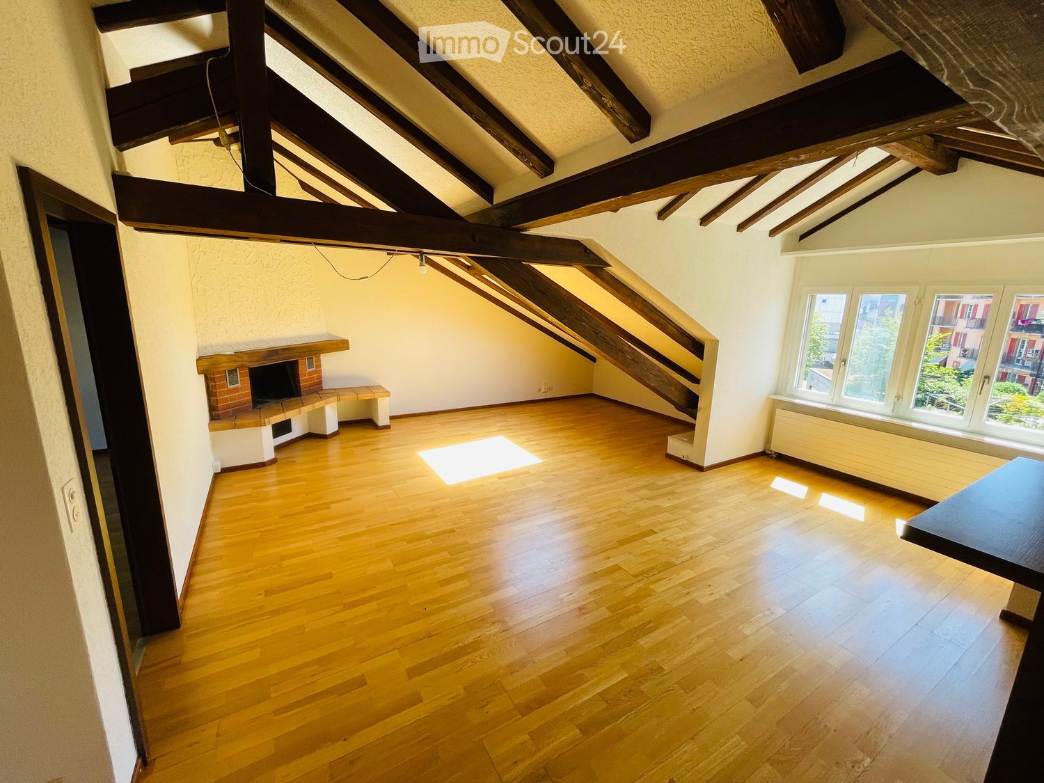 Empty room with wooden flooring, exposed wooden beams on the ceiling, a staircase leading upwards, a fireplace with a bench, and large windows letting in natural light.