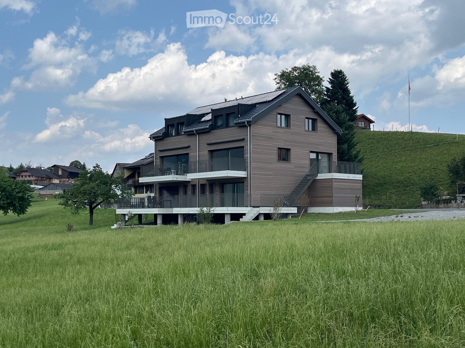 Modern two-story house with wooden siding, large windows, and balconies, situated in a grassy field with trees and a flagpole nearby.