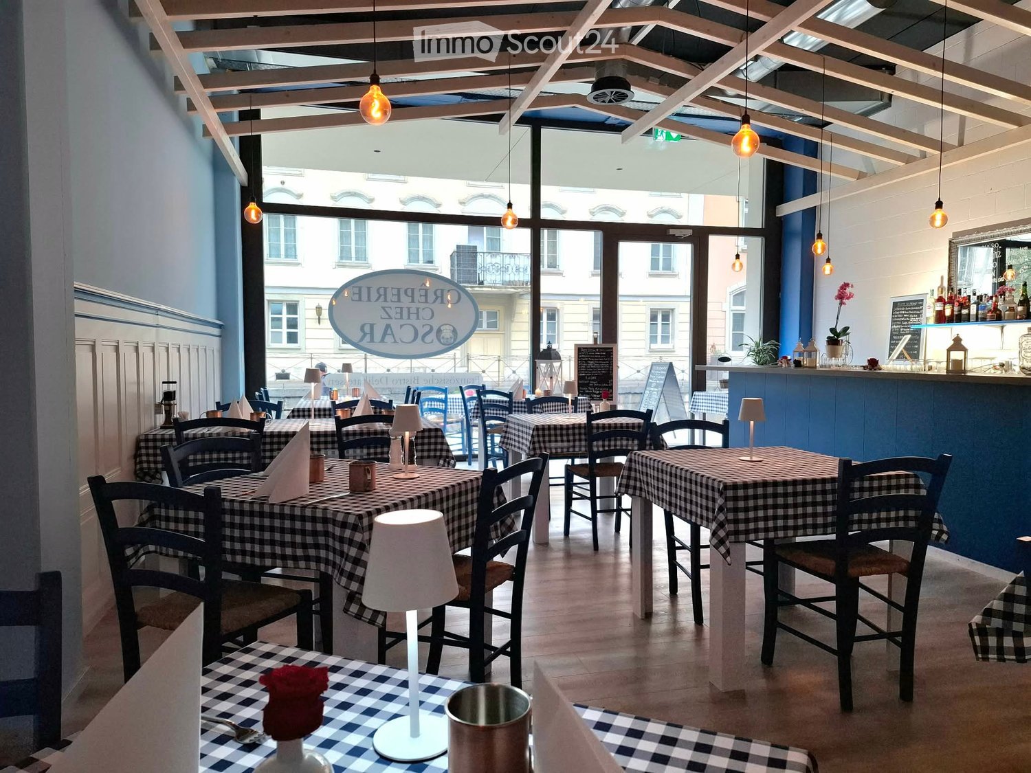 Restaurant interior with checkered tablecloths, wooden tables, chairs, pendant lights, and a bar area with bottles and a flower vase.