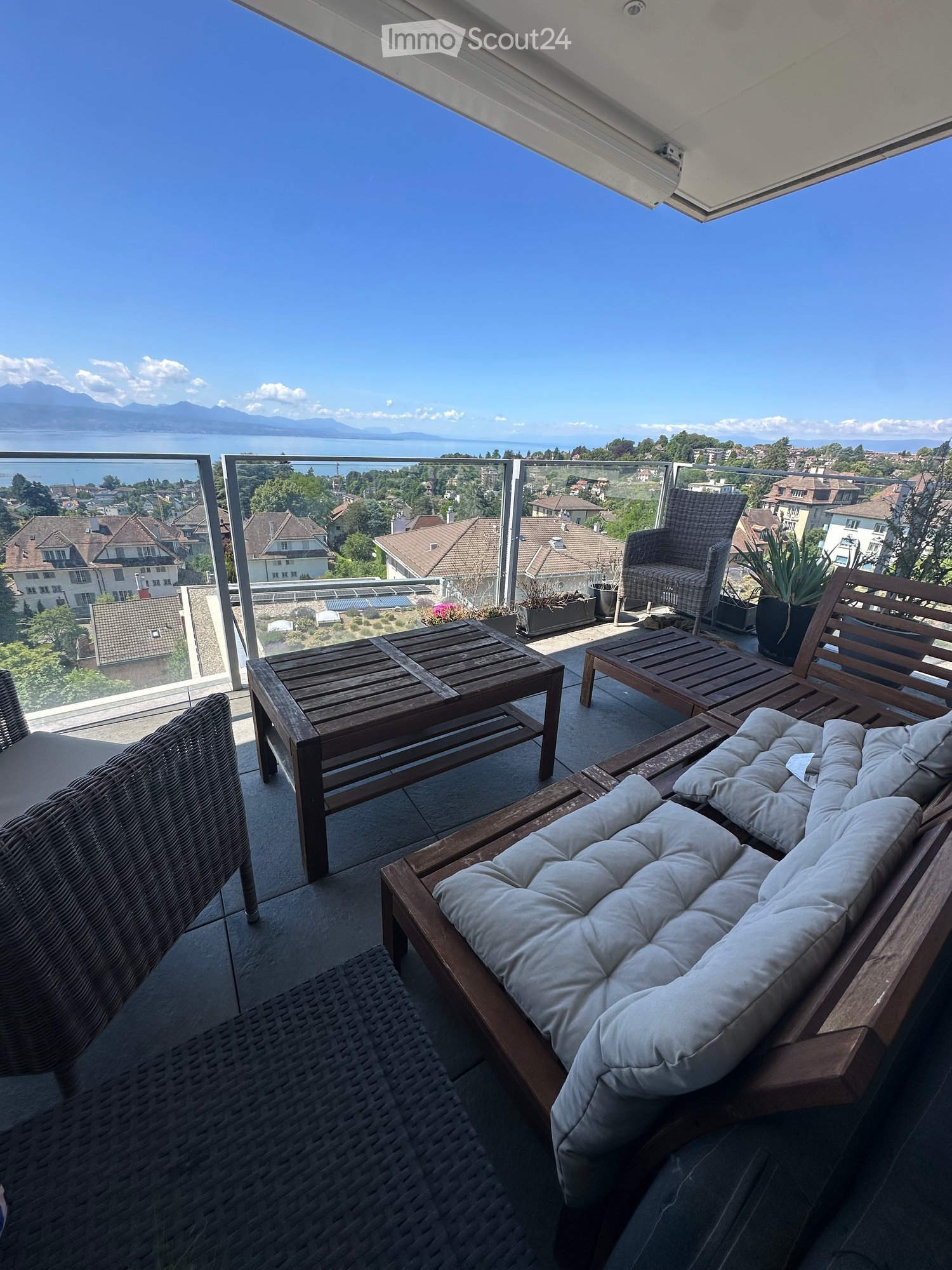 Balcony with wooden furniture, including a sofa, table, and chairs, overlooking a scenic lake and cityscape.