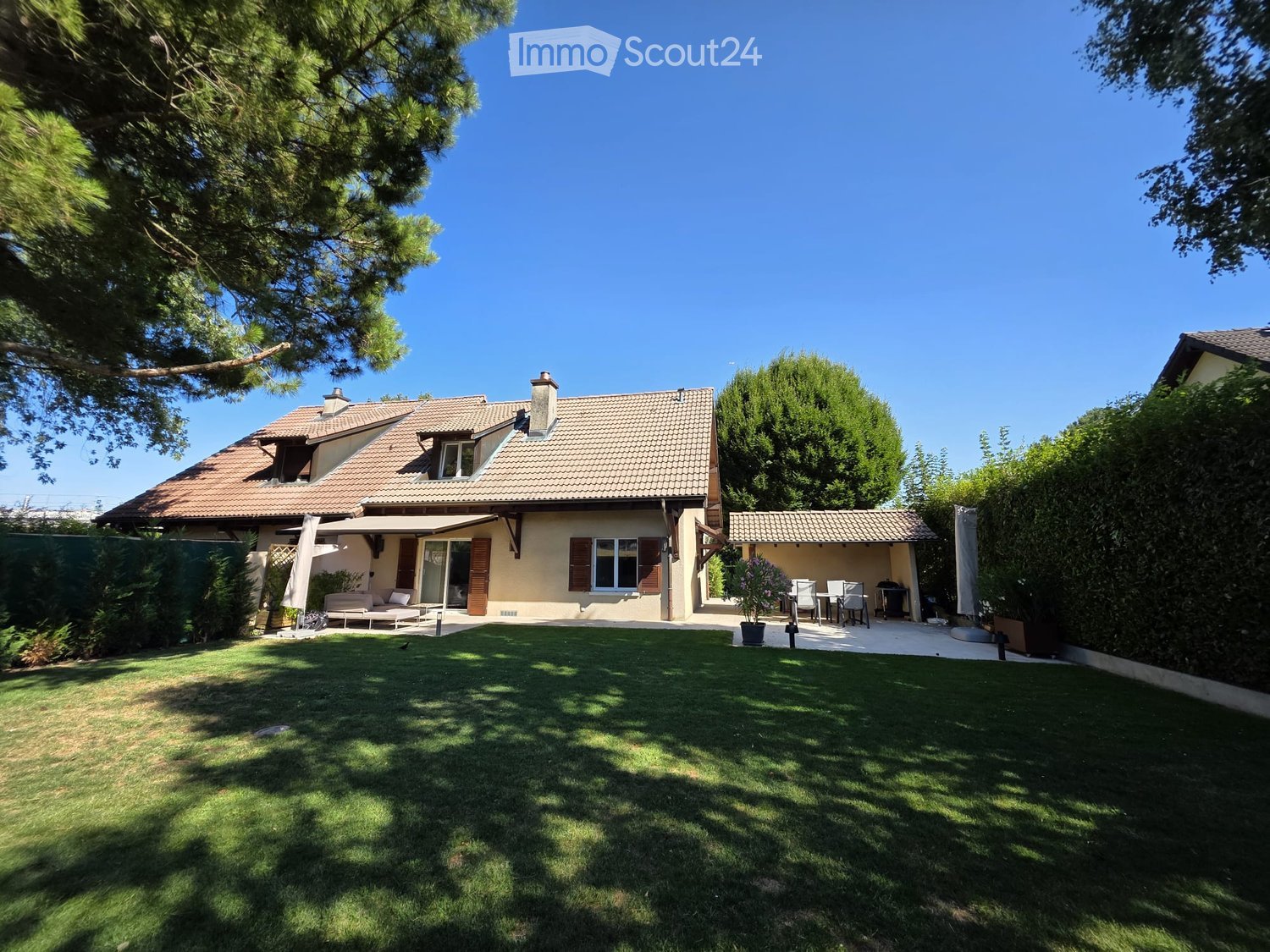 Two-story house with a tiled roof, surrounded by a well-maintained lawn and trees. Features include a covered patio area with outdoor furniture and a barbecue.