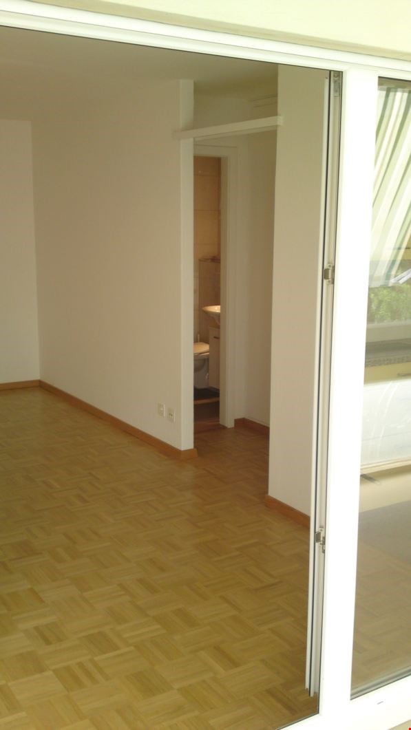 Empty room with wooden flooring and white walls, featuring a glass door leading to a balcony and an adjacent bathroom visible in the background.
