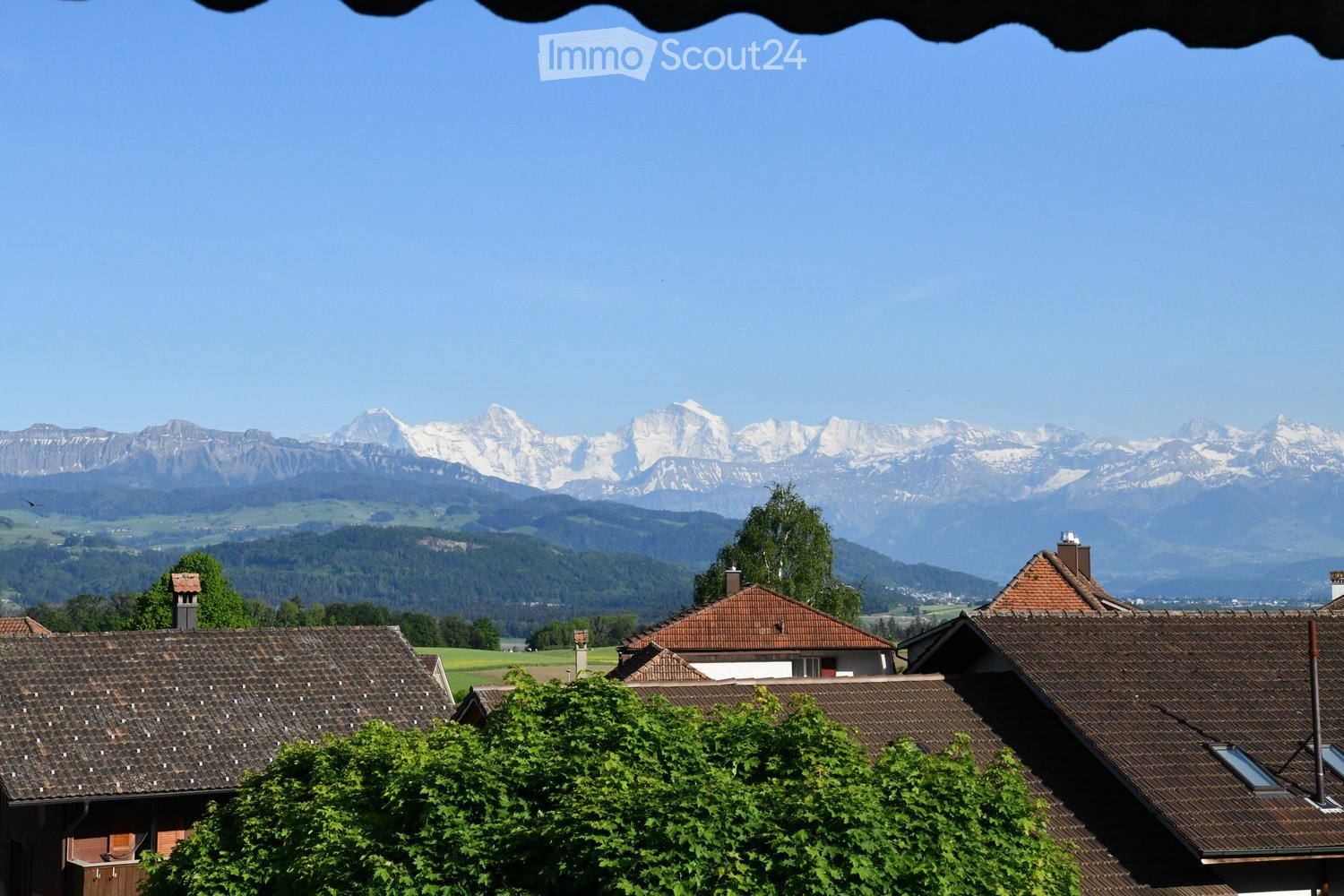 A picturesque village scene with traditional houses and red-tiled roofs, surrounded by lush green trees and a panoramic view of snow-capped mountains in the background.