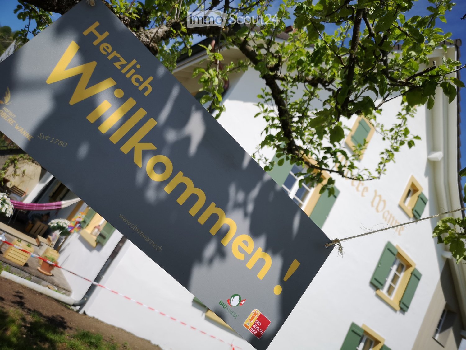 A building with white walls and green shutters, partially obscured by a sign reading "Herzlich Willkommen!" with additional logos and text.