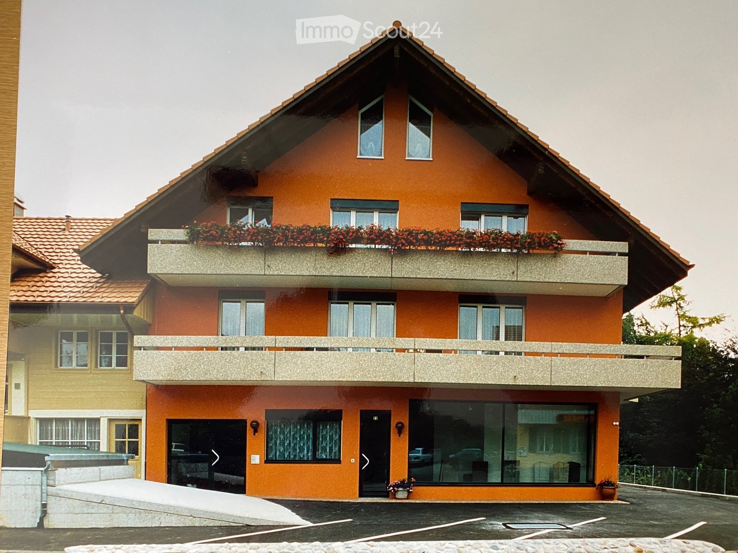 Two-story orange building with a triangular roof, multiple windows, and balconies adorned with red flowers. The ground floor features large glass windows and a black entrance door.