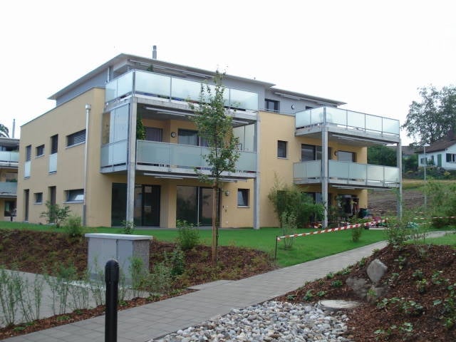 Modern two-story building with yellow and white facade, large glass balconies, and landscaped surroundings.