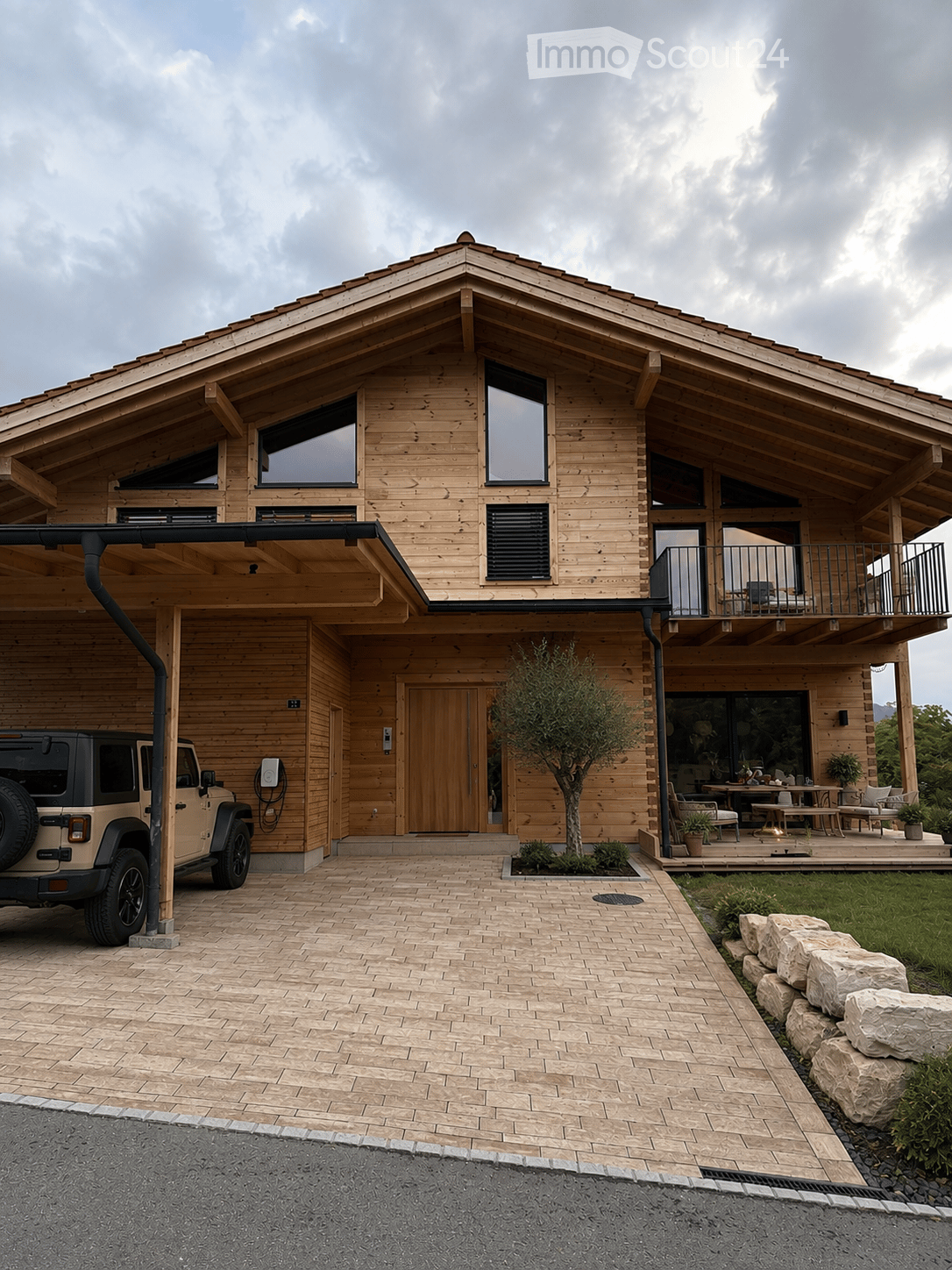Two-story wooden house with a garage, balcony, and outdoor seating area. Features large windows, a stone pathway, and a charging station for an electric vehicle.