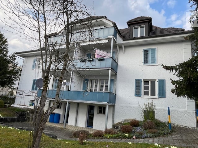 A three-story white building with blue shutters and balconies, featuring a raised ground floor and a small garden area.