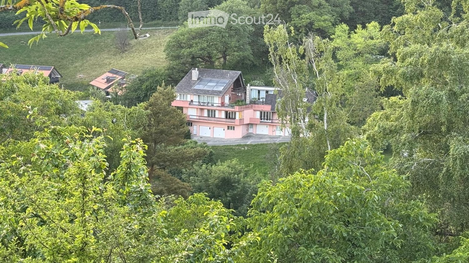 A two-story house with a pink exterior, white garage doors, and a solar panel on the roof, surrounded by lush greenery and trees.