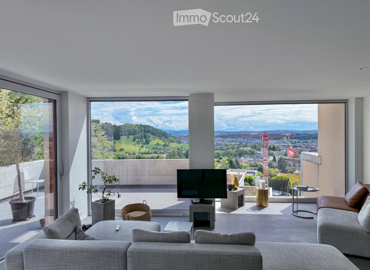Modern living room with large windows offering a scenic view, featuring a grey sofa, coffee table, armchair, television, small table, and potted plants.