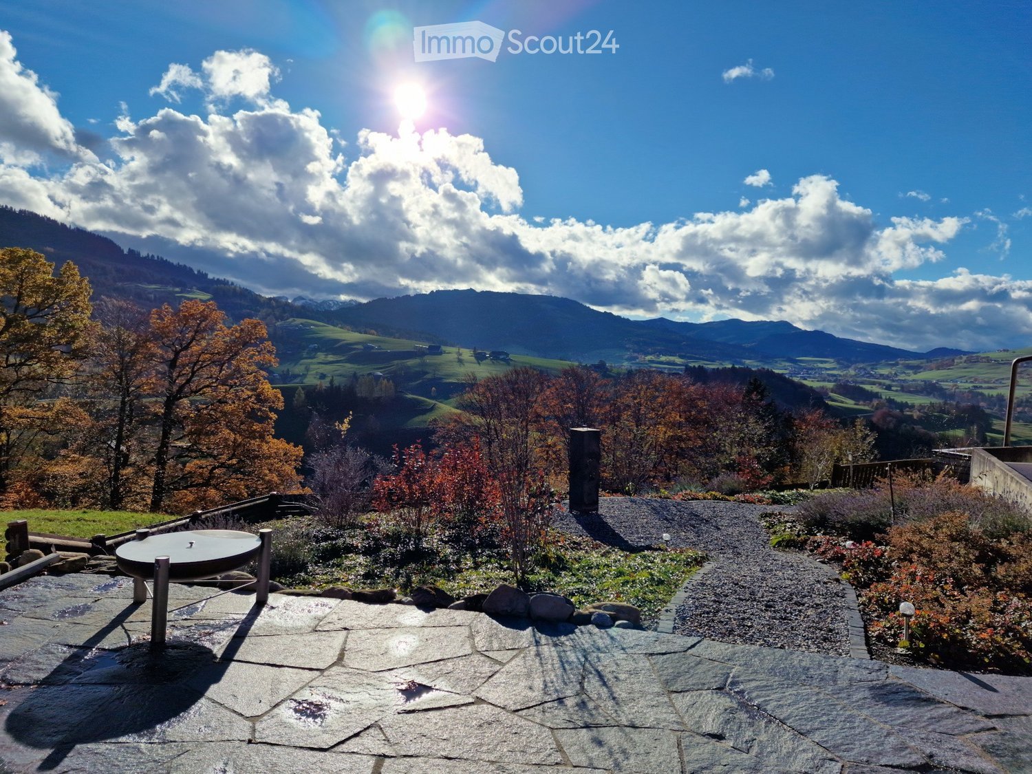 A serene outdoor garden area with a stone-paved patio, a round metal table, and a scenic mountain view in the background.