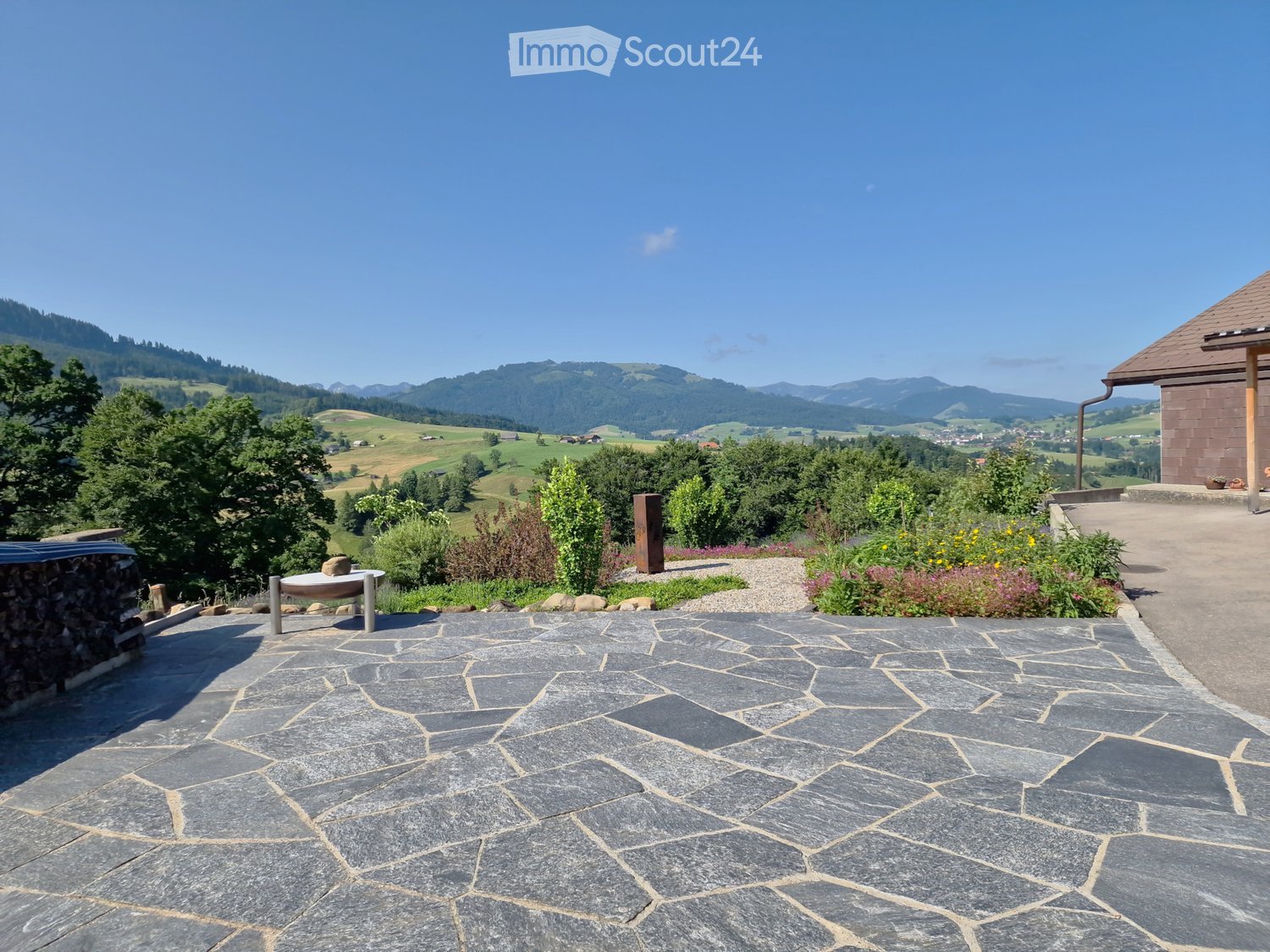 A spacious outdoor area with a stone-paved patio, surrounded by greenery and a scenic mountain view in the background.