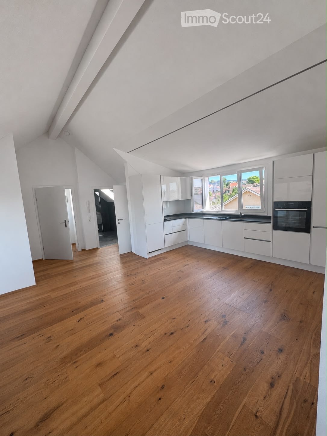 Empty kitchen with white cabinets, wooden flooring, and large windows offering a view of the outside. The space is clean and unfurnished, with no appliances or furniture present.