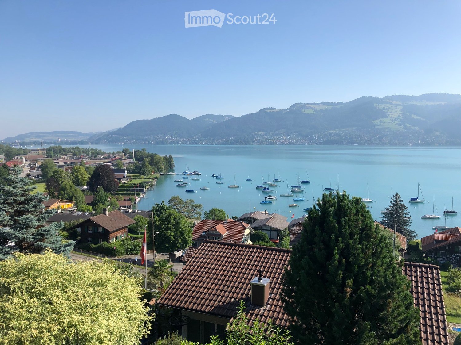A scenic view of a lakeside town with numerous sailboats docked in the water, surrounded by green trees and houses with red-tiled roofs. The background features a calm lake and distant mountains.