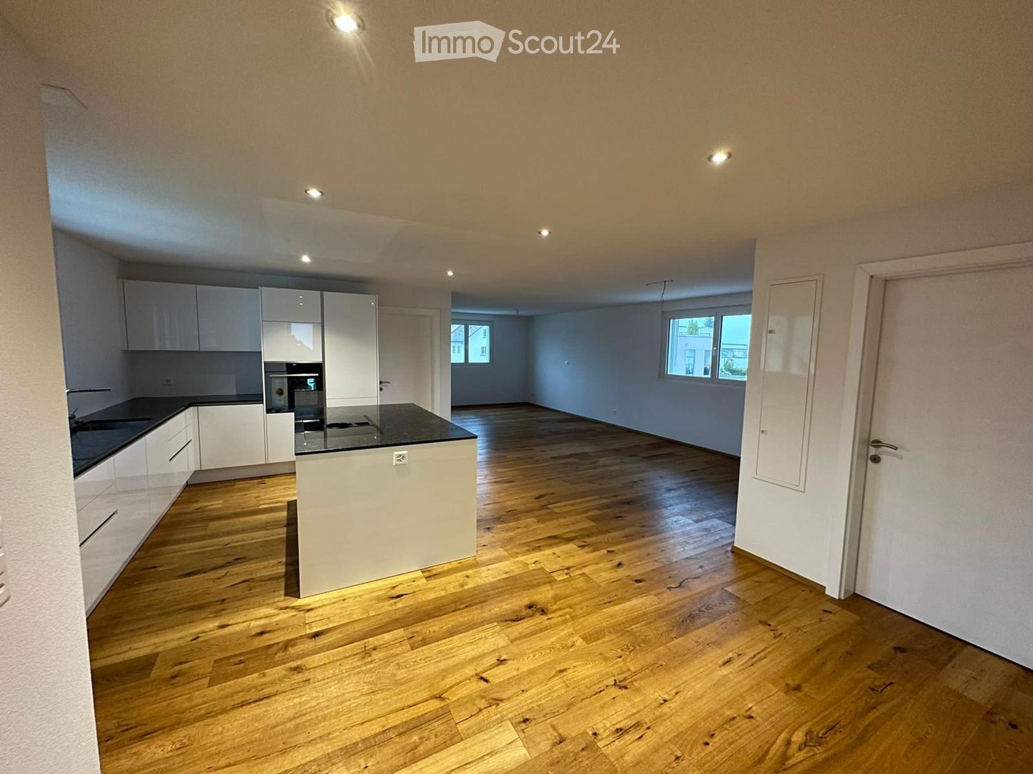 Modern kitchen with white cabinets, a central island, and wooden flooring. The space is open and well-lit with recessed lighting.