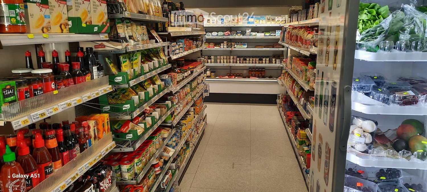 A grocery store aisle with shelves stocked with various food products, including sauces, condiments, and packaged goods. A refrigerator unit is visible on the right side with fresh produce inside.