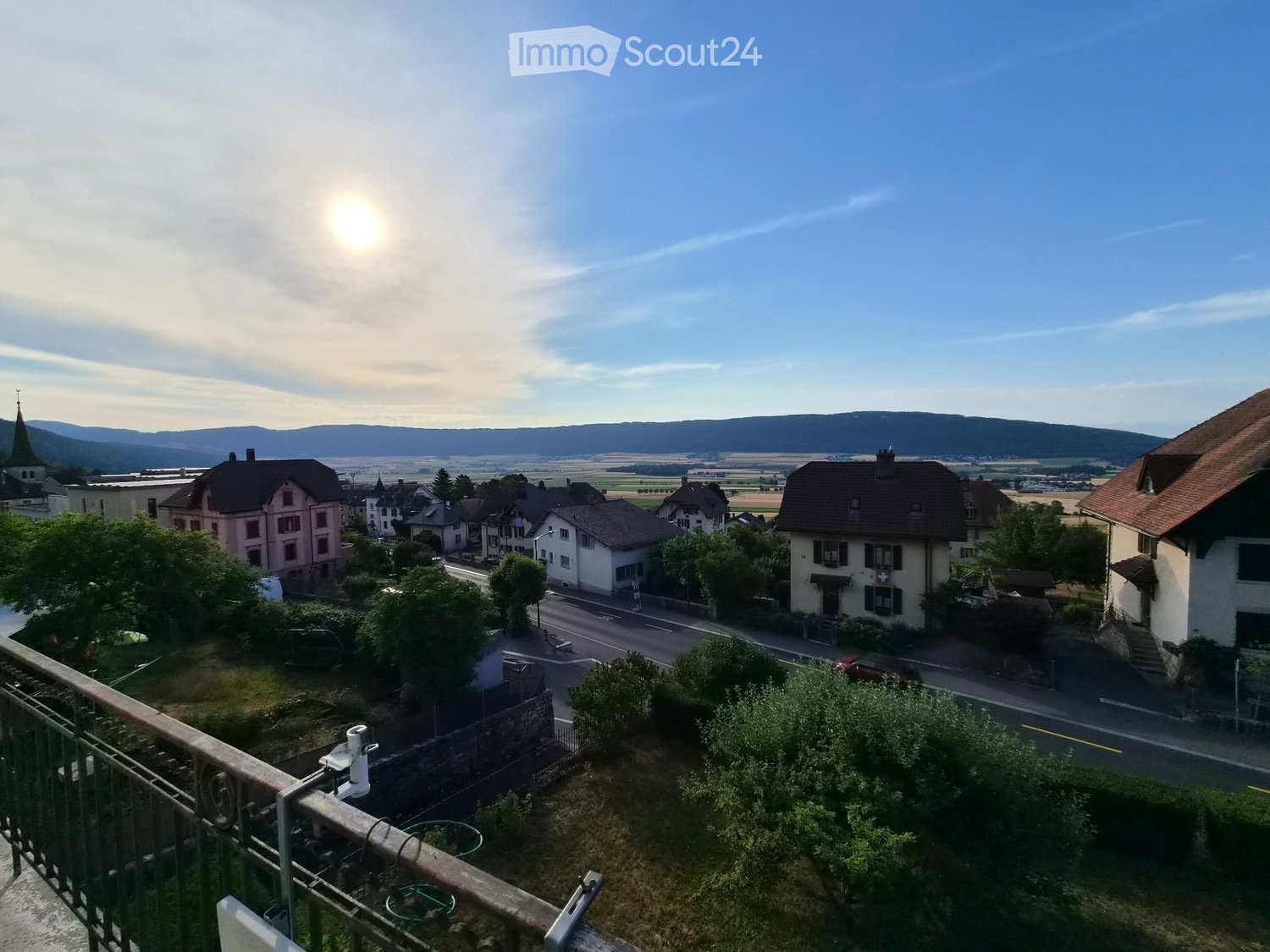 Balcony overlooking a small town with houses, a road, and distant hills under a clear sky.