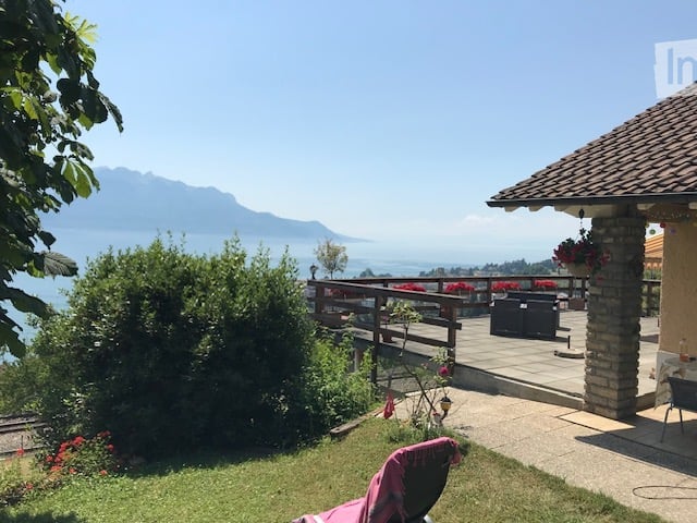 Garden with a view of a lake and mountains, featuring a wooden deck, potted plants, and outdoor furniture.