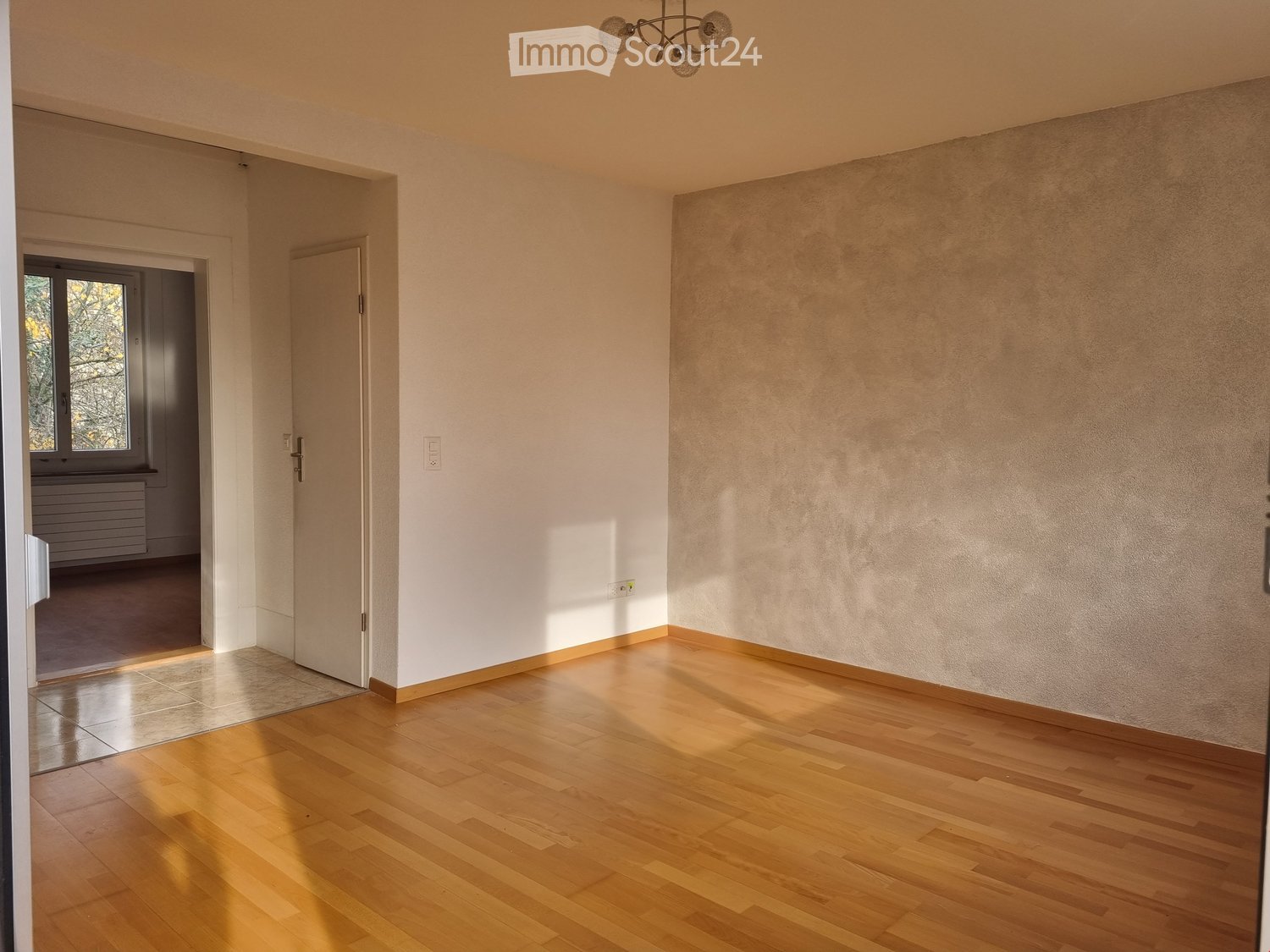 Empty room with wooden flooring, white walls, and a door leading to another room. Natural light streams in from a window in the adjacent room.