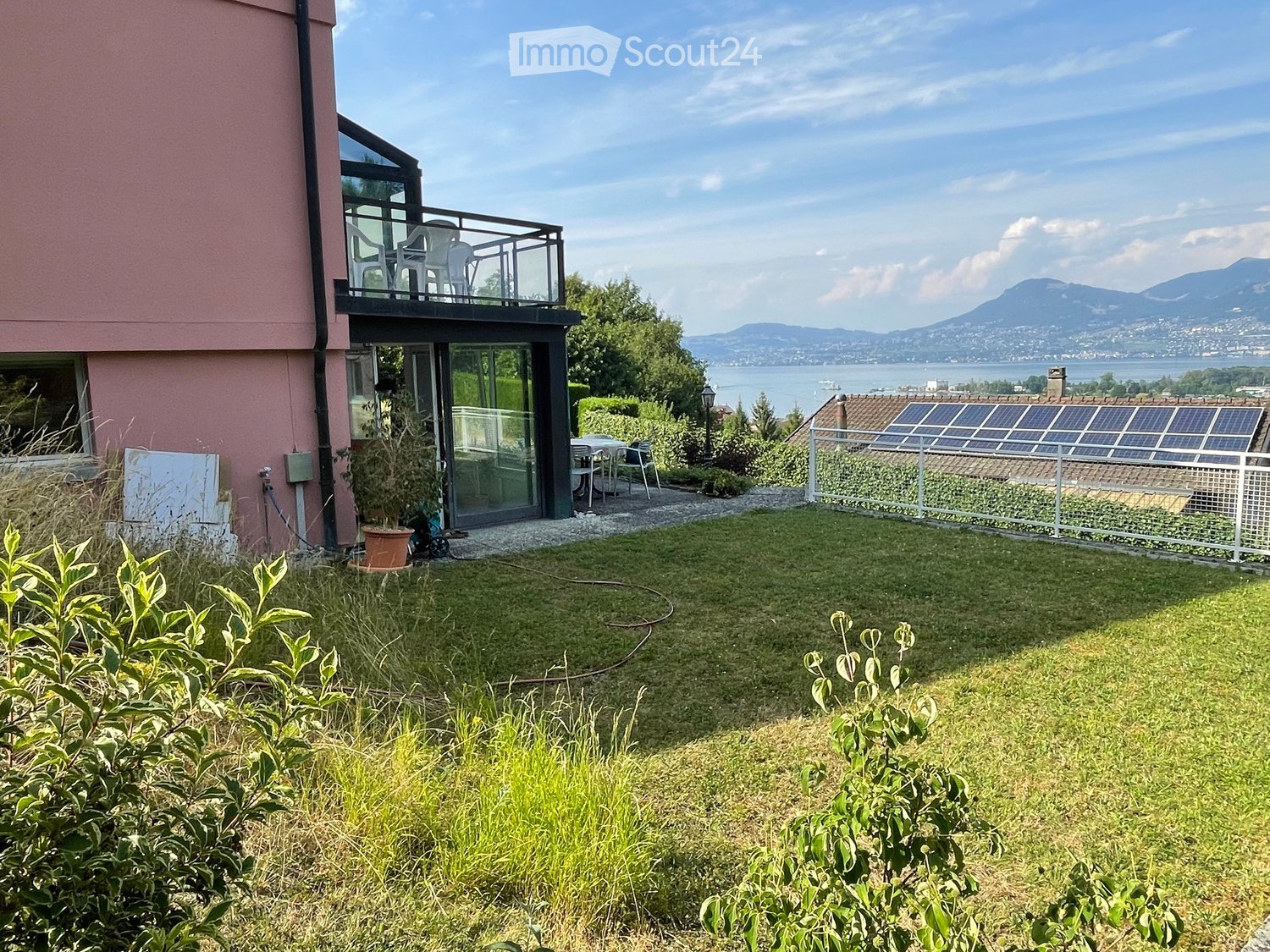 Garden with lawn, patio furniture, balcony with chairs, solar panels on neighboring roof, scenic lake and mountain view.