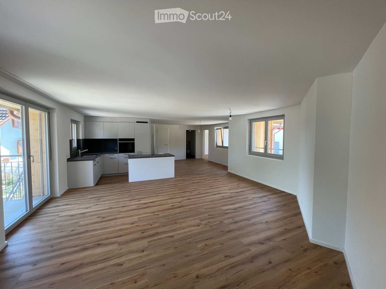 Empty room with wooden flooring, white walls, and a small kitchen area with cabinets and a sink visible in the background.