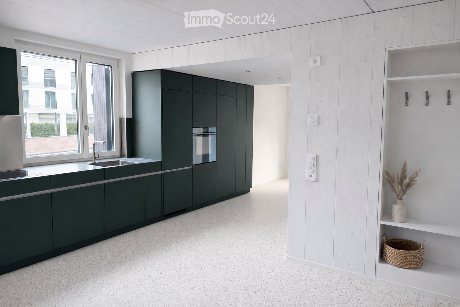 Modern kitchen with dark green cabinets, white wooden walls, and a white tiled floor. Features a sink with a modern faucet, built-in oven, and open shelving with decorative items.