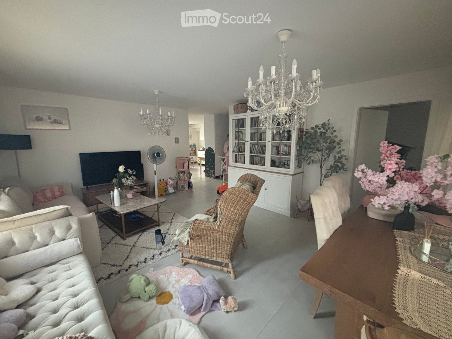 A well-lit living room with a cream sofa, coffee table, wicker chair, dining table, and decorative items including a chandelier and cabinet.