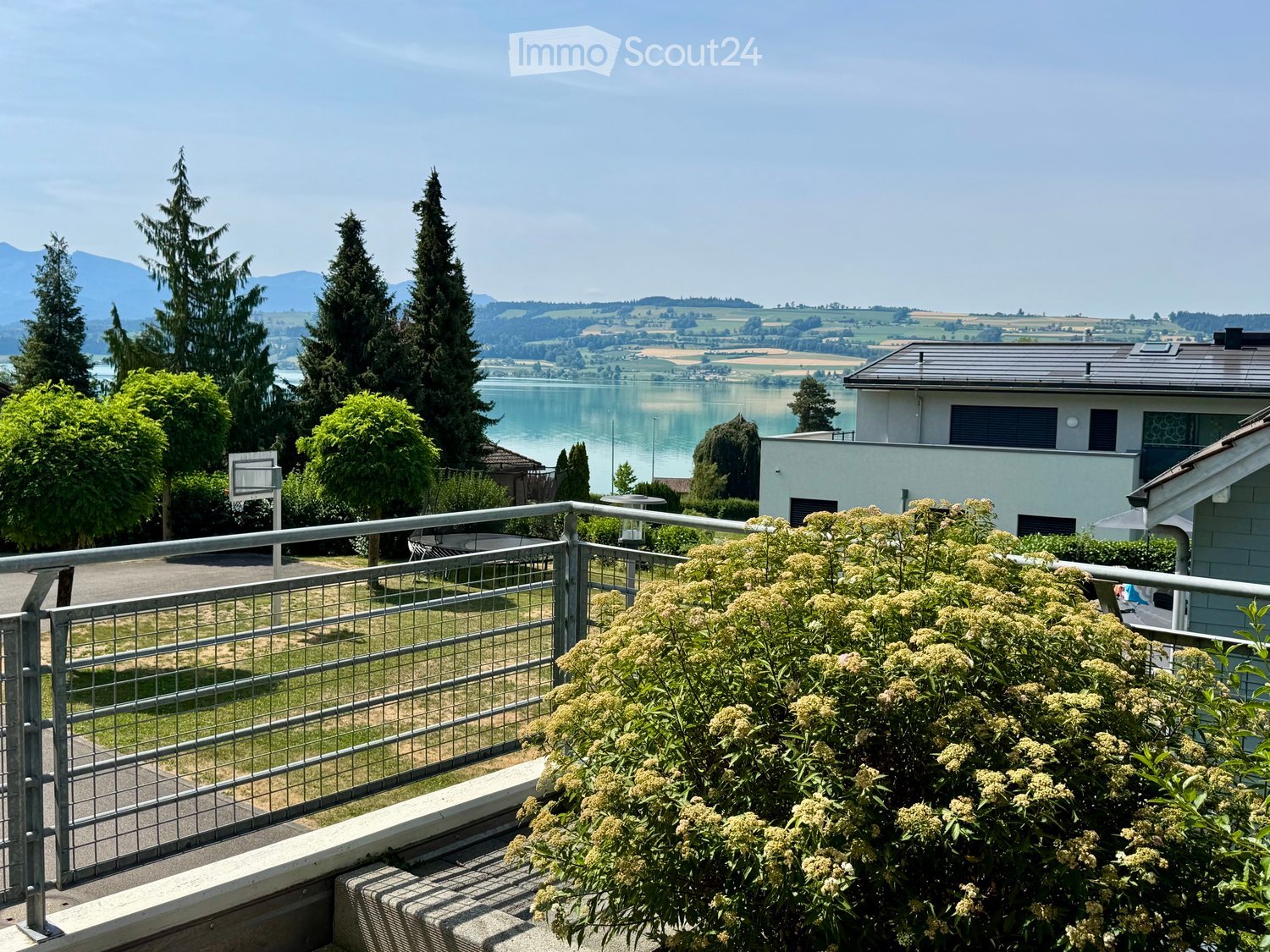 Balcony with metal railing, lush greenery, and scenic view of a lake and mountains in the background.
