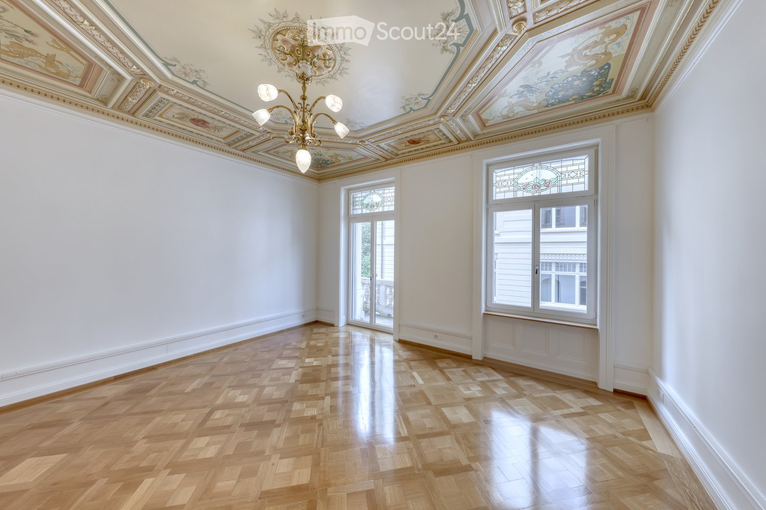 An empty room with polished wooden flooring, white walls, and ornate ceiling details. Two large windows with decorative glass panels and a chandelier hanging from the ceiling.