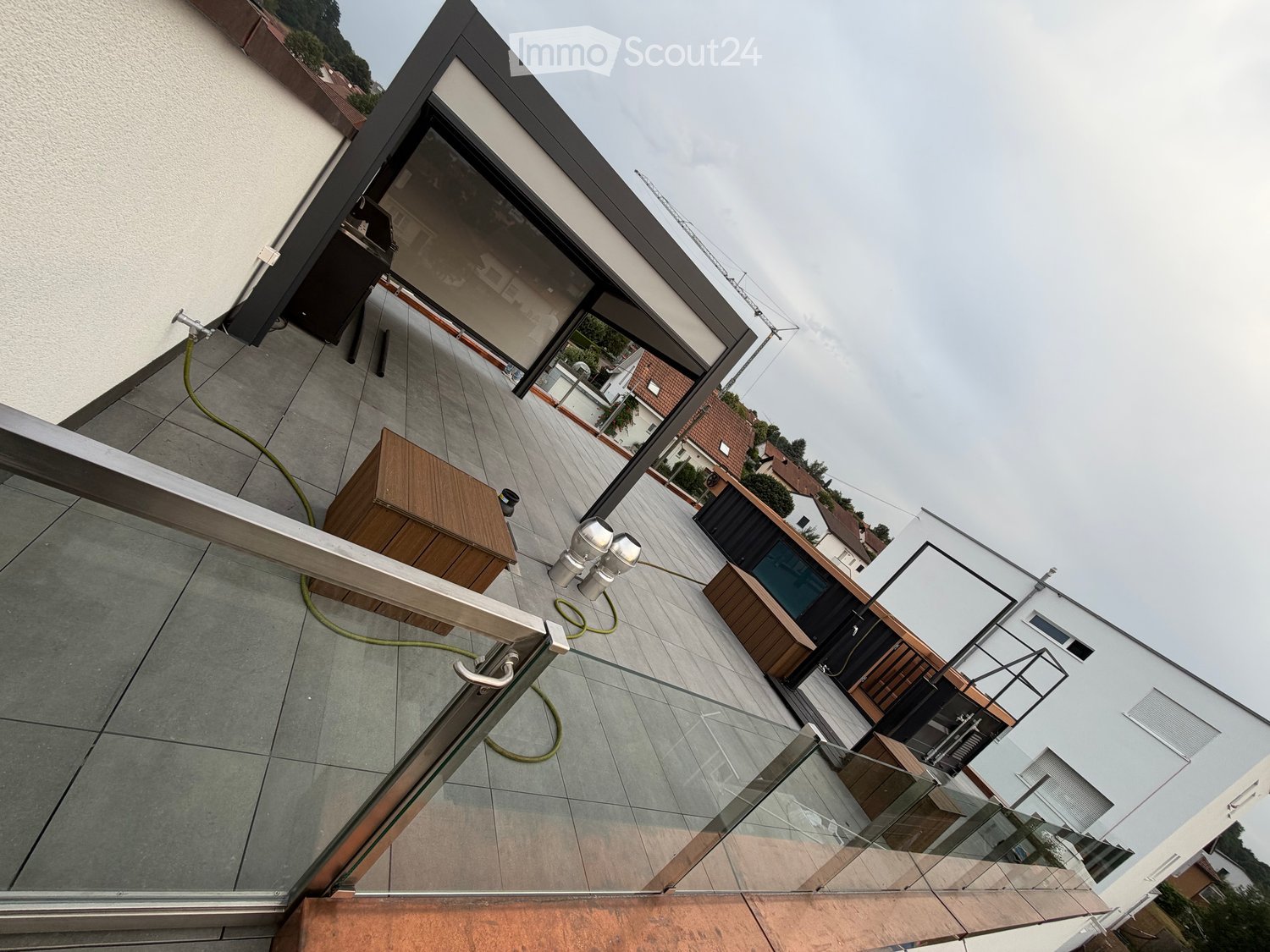 Balcony with modern glass railing, tiled floor, and wooden furniture, overlooking a residential area with neighboring houses and greenery.