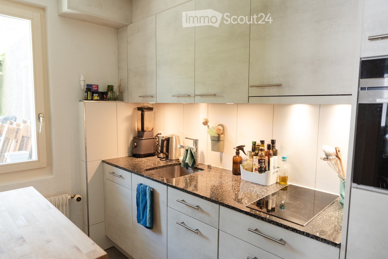 Modern kitchen with white cabinets, granite countertop, induction stove, blender, electric kettle, and various bottles and items on the counter.