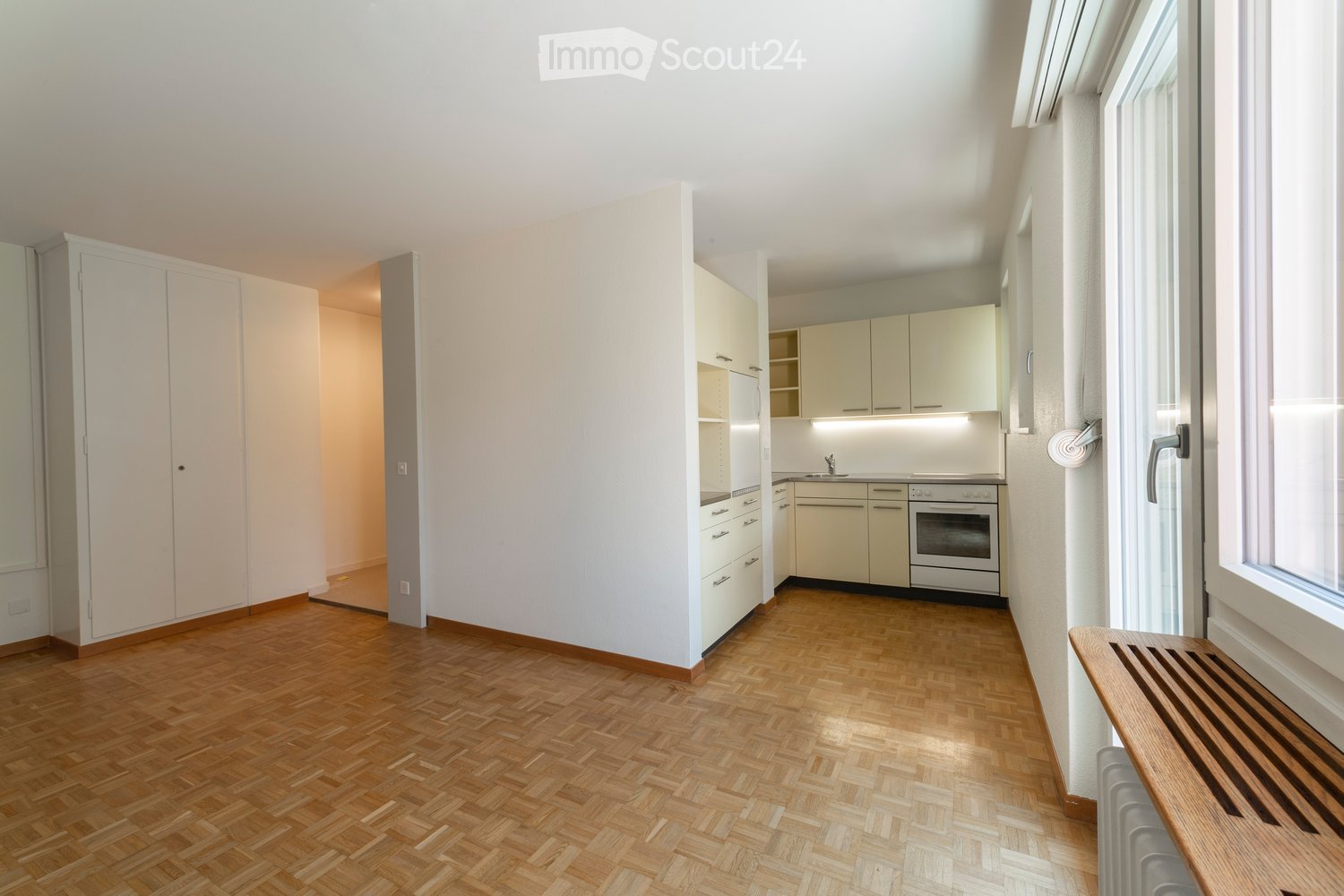 A modern, unfurnished kitchen with light-colored cabinets, a refrigerator, an oven, and a sink. The floor is made of herringbone-patterned wood.