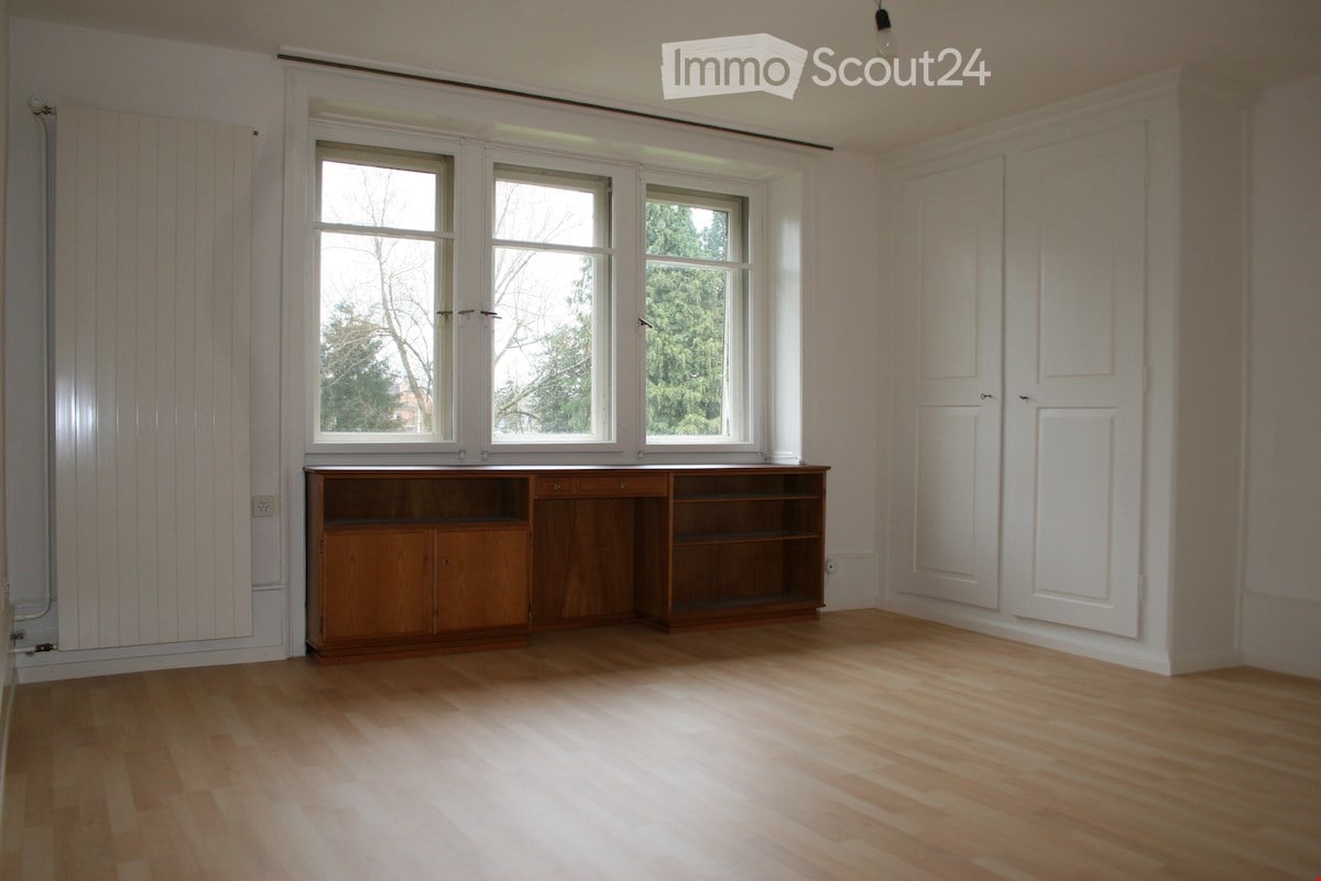 Empty room with wooden flooring, white walls, large windows, and a wooden cabinet unit against one wall.