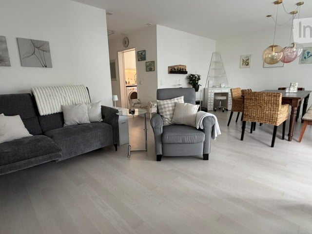 Modern living room with light wood flooring, grey sofa, armchair, coffee table, dining area with chairs, and decorative wall art.