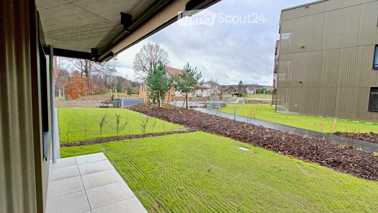 Well-maintained garden area with neatly trimmed grass, mulch beds, small trees, and a paved pathway leading to a modern building.