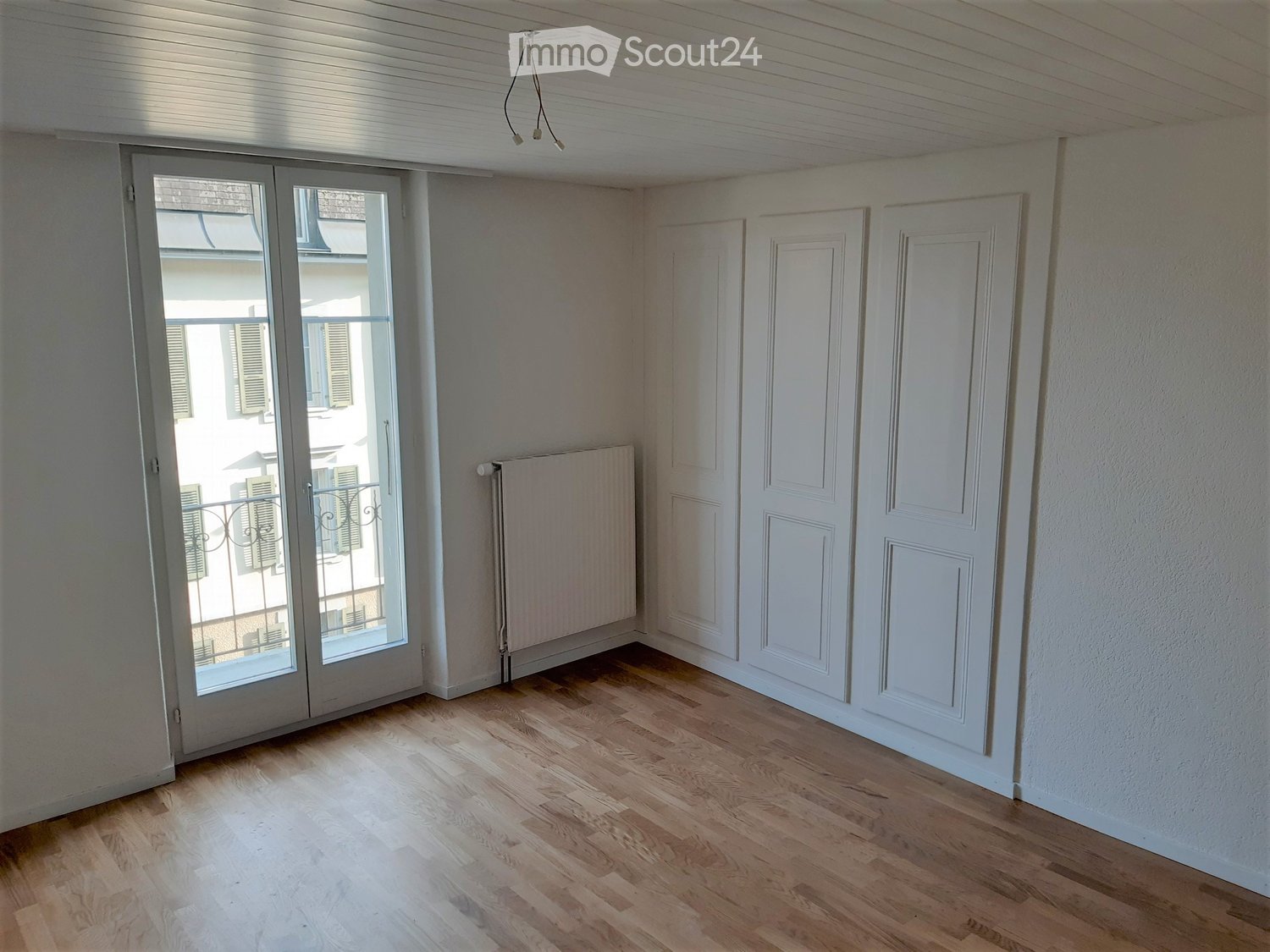Empty room with wooden flooring, white walls, and a large window with a view of a balcony and outdoor area.