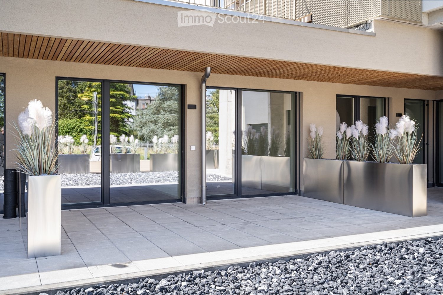 Modern outdoor area with large glass sliding doors, tiled floor, and tall metallic planters with white decorative plants.
