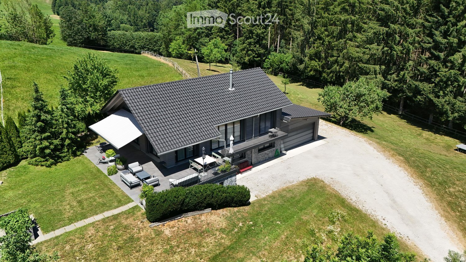 A modern two-story house with a grey exterior, dark roof, and large windows, situated on a grassy plot with a driveway, patio, and outdoor seating area.