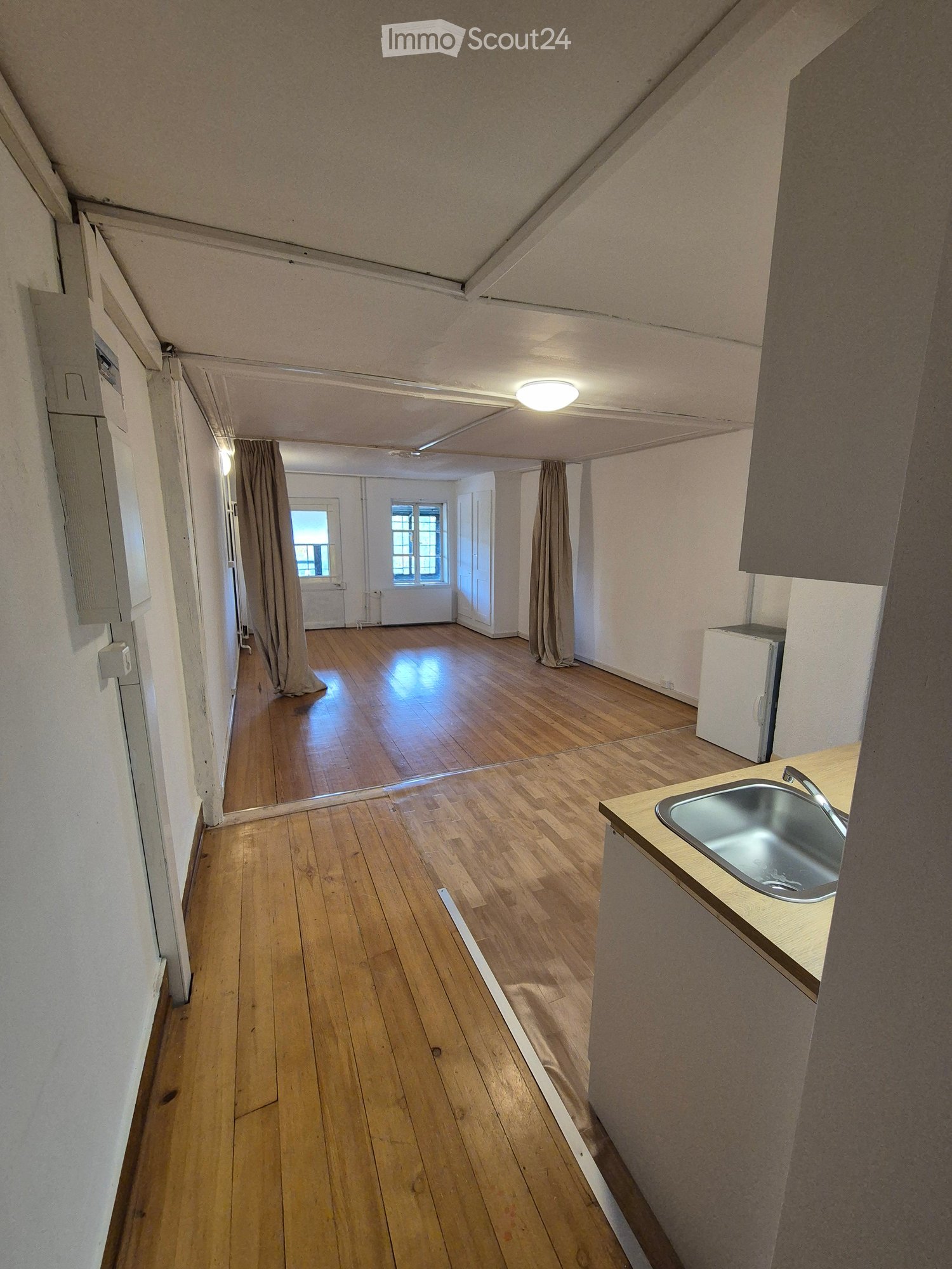 A narrow hallway with wooden flooring, leading to a small kitchen area with a sink and a refrigerator. The walls are painted white, and there are curtains on the windows at the end of the hallway.