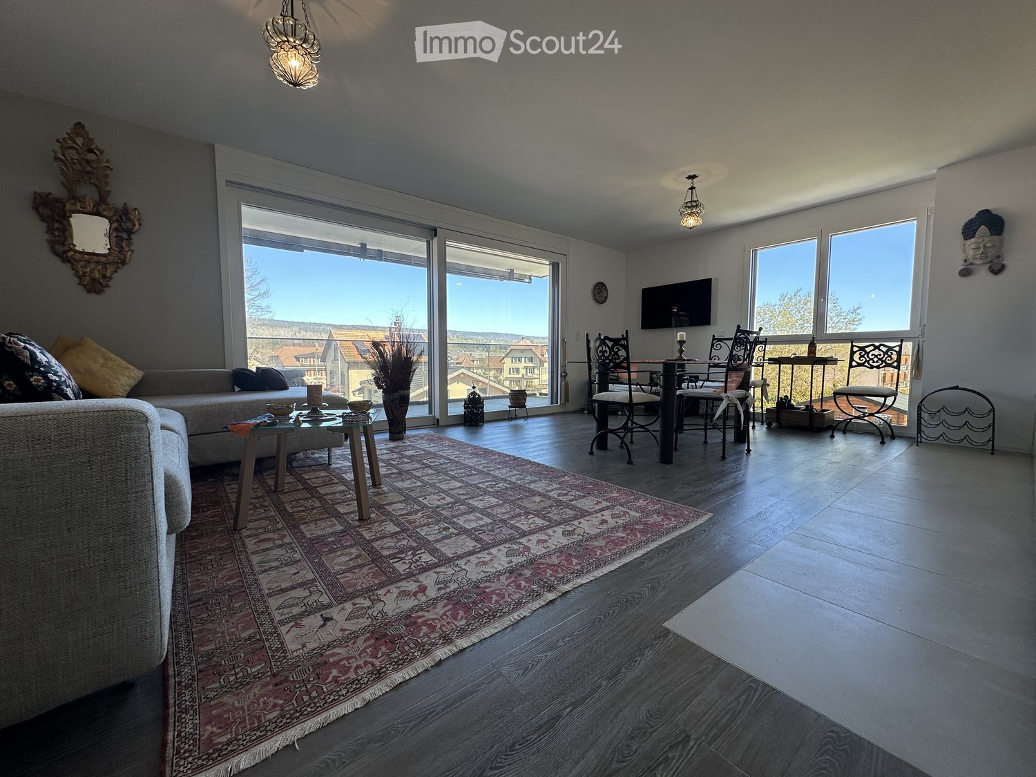 Bright living room with large windows offering a scenic view, featuring a sofa, coffee table, dining area with chairs, and decorative elements like a mirror and vase.