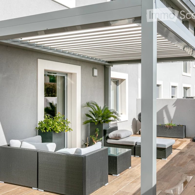Modern outdoor patio with a covered pergola, wicker sofa, glass coffee table, lounge chairs, and potted plants.