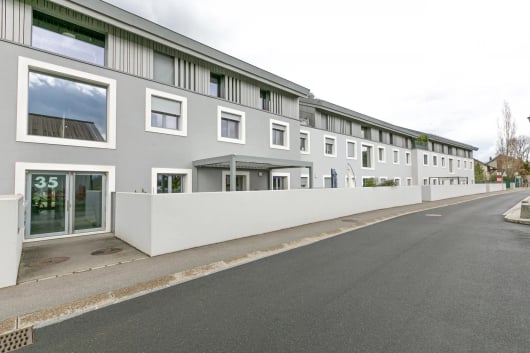Modern multi-unit residential building with grey facade, white-framed windows, and a white privacy wall along the street.