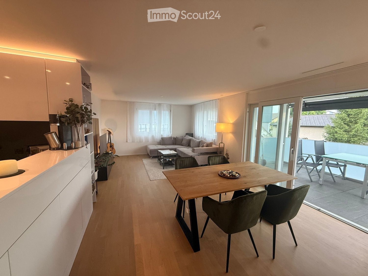 Open-plan living and dining area with light wood flooring, white walls, large windows, and a sliding glass door leading to a balcony. Features a grey sectional sofa, coffee table, dining table with chairs, and a kitchen counter with a vase and plant.
