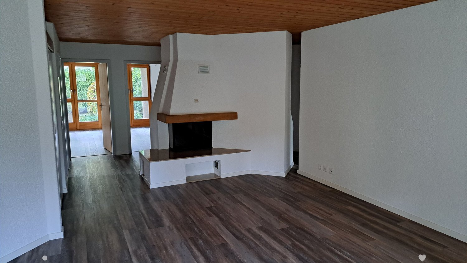 Empty living room with wooden flooring, white walls, and a built-in fireplace. Two large glass doors lead to an outdoor area.