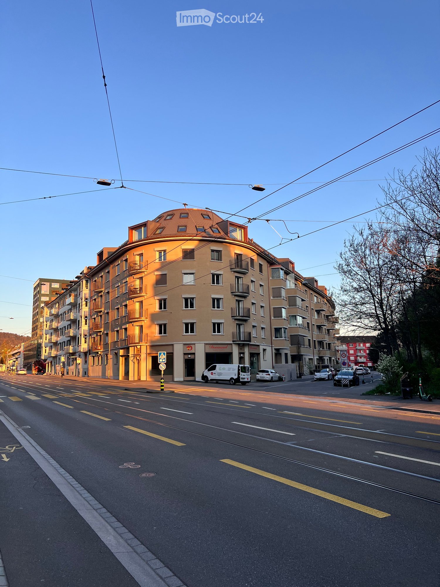 Multi-story building with a rounded facade, balconies, and large windows, located on a street with parked cars and traffic markings.