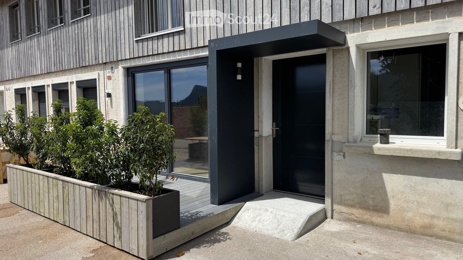 Modern outdoor area with raised wooden planter box containing small trees, glass sliding doors, and a sleek black entrance door.