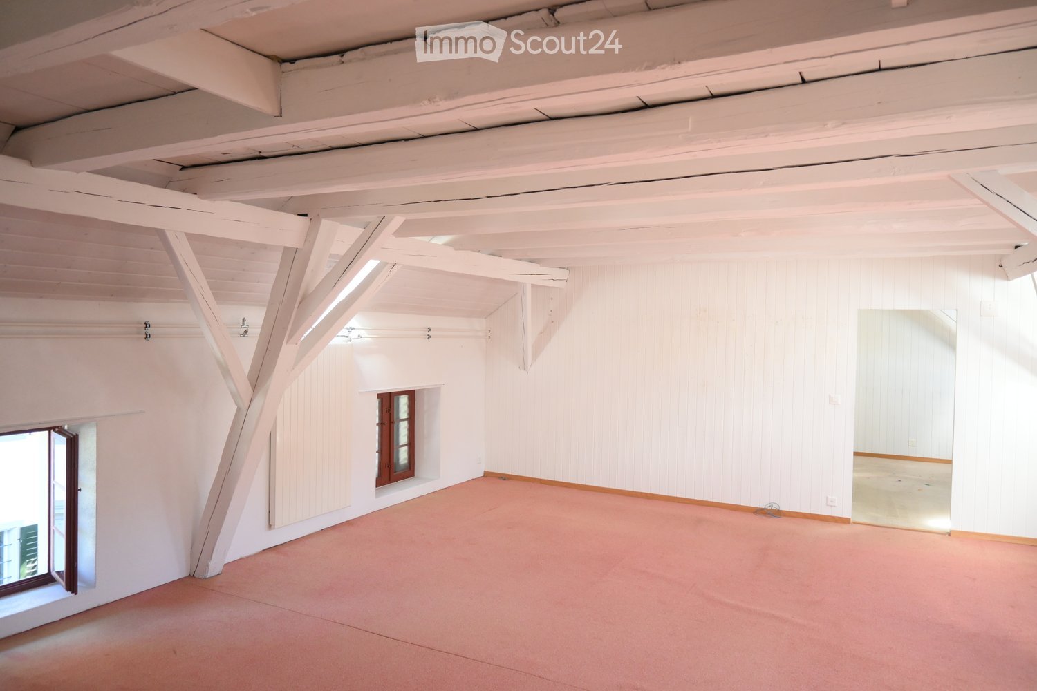 Empty room with white walls, exposed wooden beams, and a pink carpeted floor. Features include a large window on the left and a smaller windowed doorway in the center.