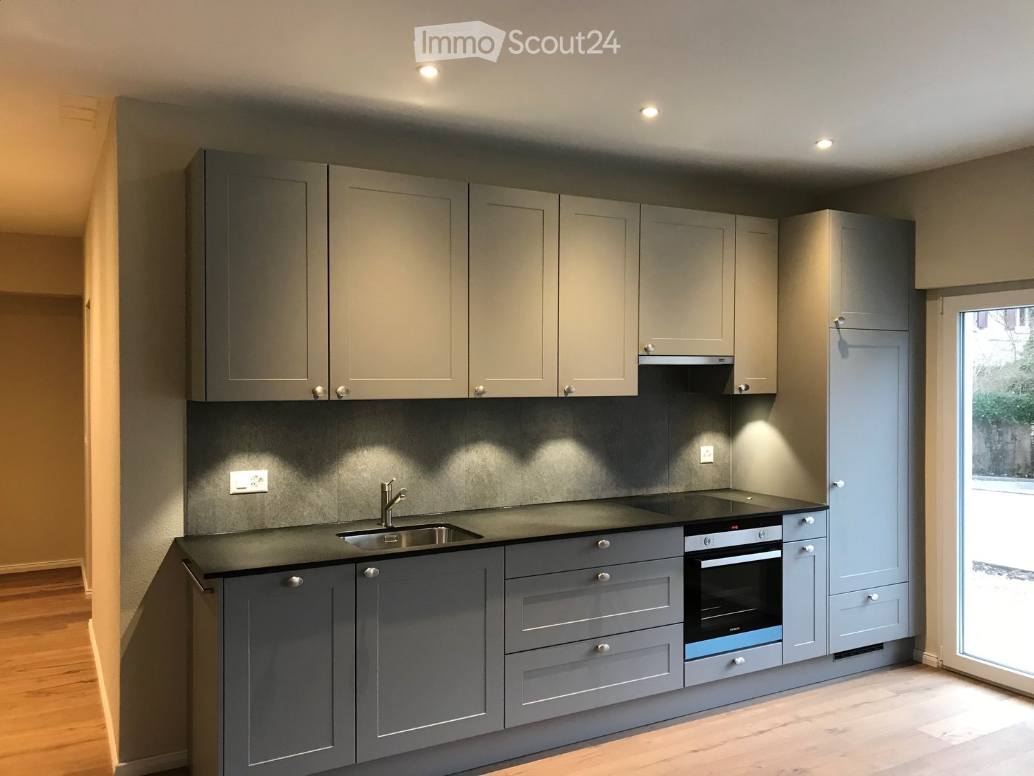 Modern kitchen with grey cabinets, black countertop, stainless steel sink, and built-in oven. Features recessed lighting and a large window.
