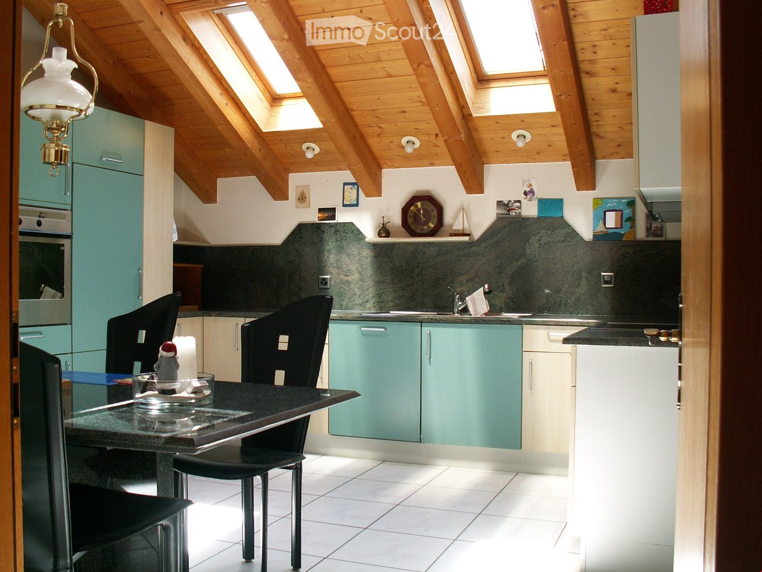 Wooden ceiling, wooden floor, green cupboards, white kitchen table, black chairs, countertop, kitchen appliances, wall mounted lamps