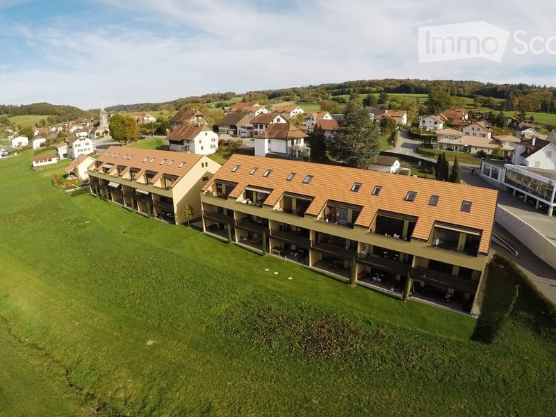 Aerial view of a modern multi-story residential building with orange-tiled roofs and balconies, surrounded by green lawns and neighboring houses in a rural setting.